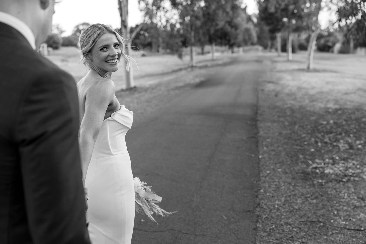 A joyful bride in a fitted strapless gown smiles while holding a bouquet on a tree-lined path at the Corowa Distillery.