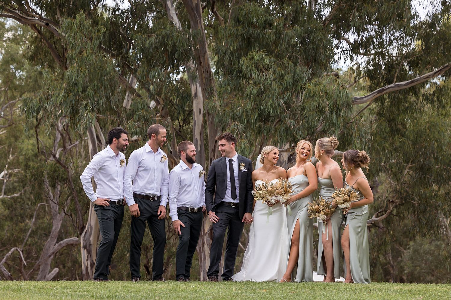 A wedding party in formal attire poses together on a grassy hill with eucalyptus trees in the background at Quandong in Wahgunyah.
