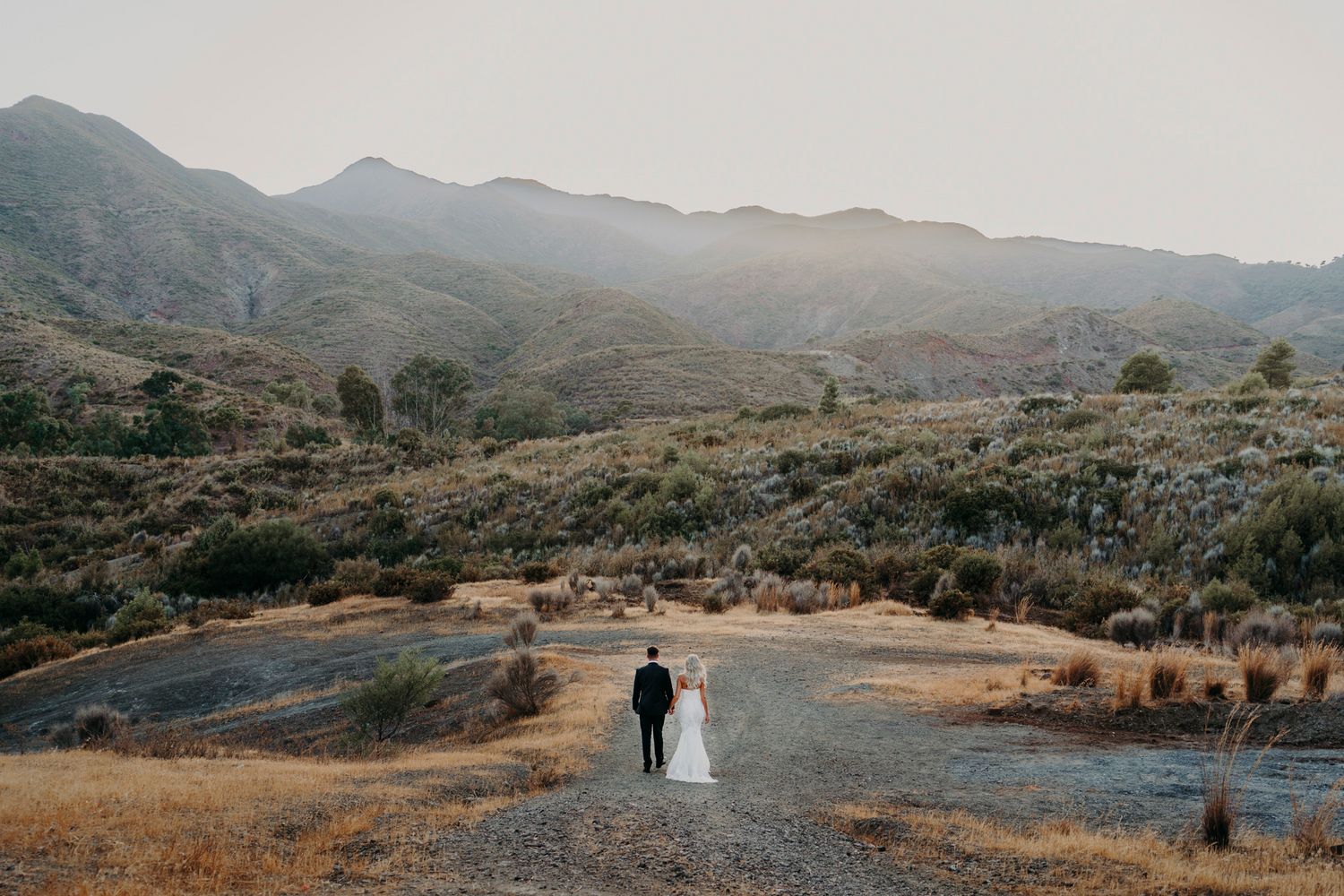 A lone figure walks on a winding dirt path through dry mountainous terrain at golden hour.