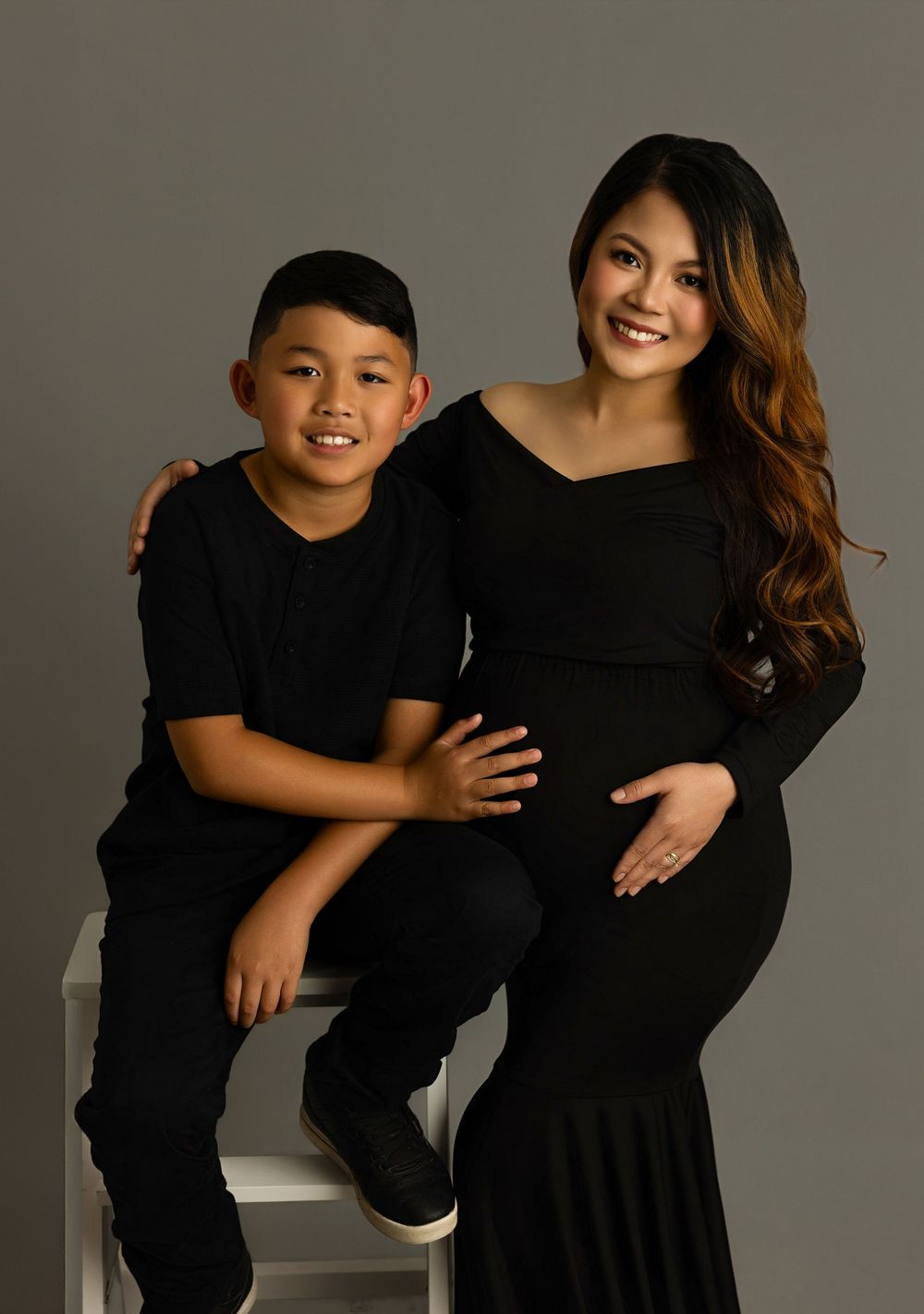A series of candid maternity photos showing a woman in black dress and young child in black shirt posing together on gray backdrop.