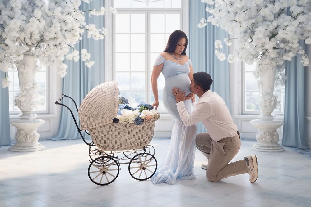 Expectant couple in elegant blue studio, man kissing pregnant woman's belly near vintage white wicker baby carriage with flowers.