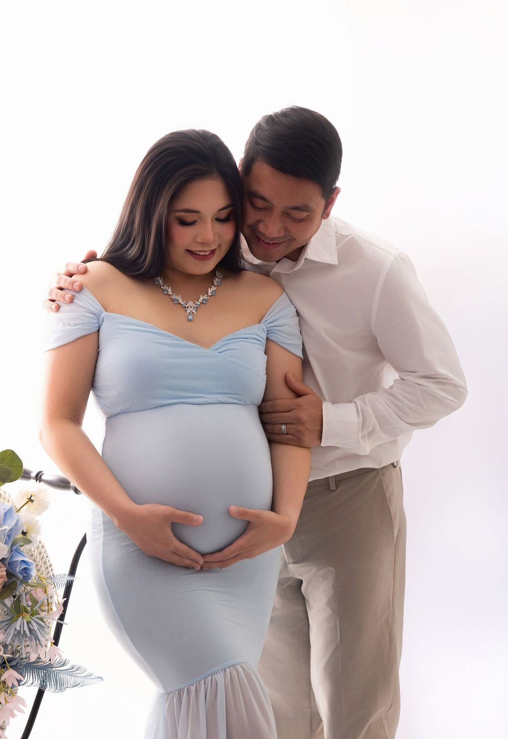 Couple in maternity photoshoot, woman in light blue gown cradling baby bump, man in white shirt embracing her, white background.