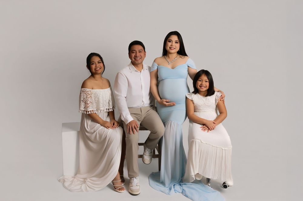 Pregnant woman in blue gown poses with family in white outfits for a maternity studio portrait.