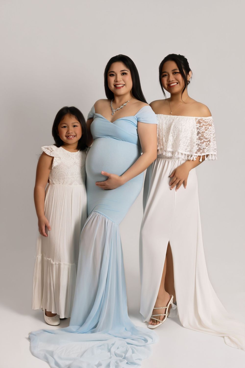 Pregnant woman in blue gown poses with two daughters in white dresses for a maternity studio portrait.