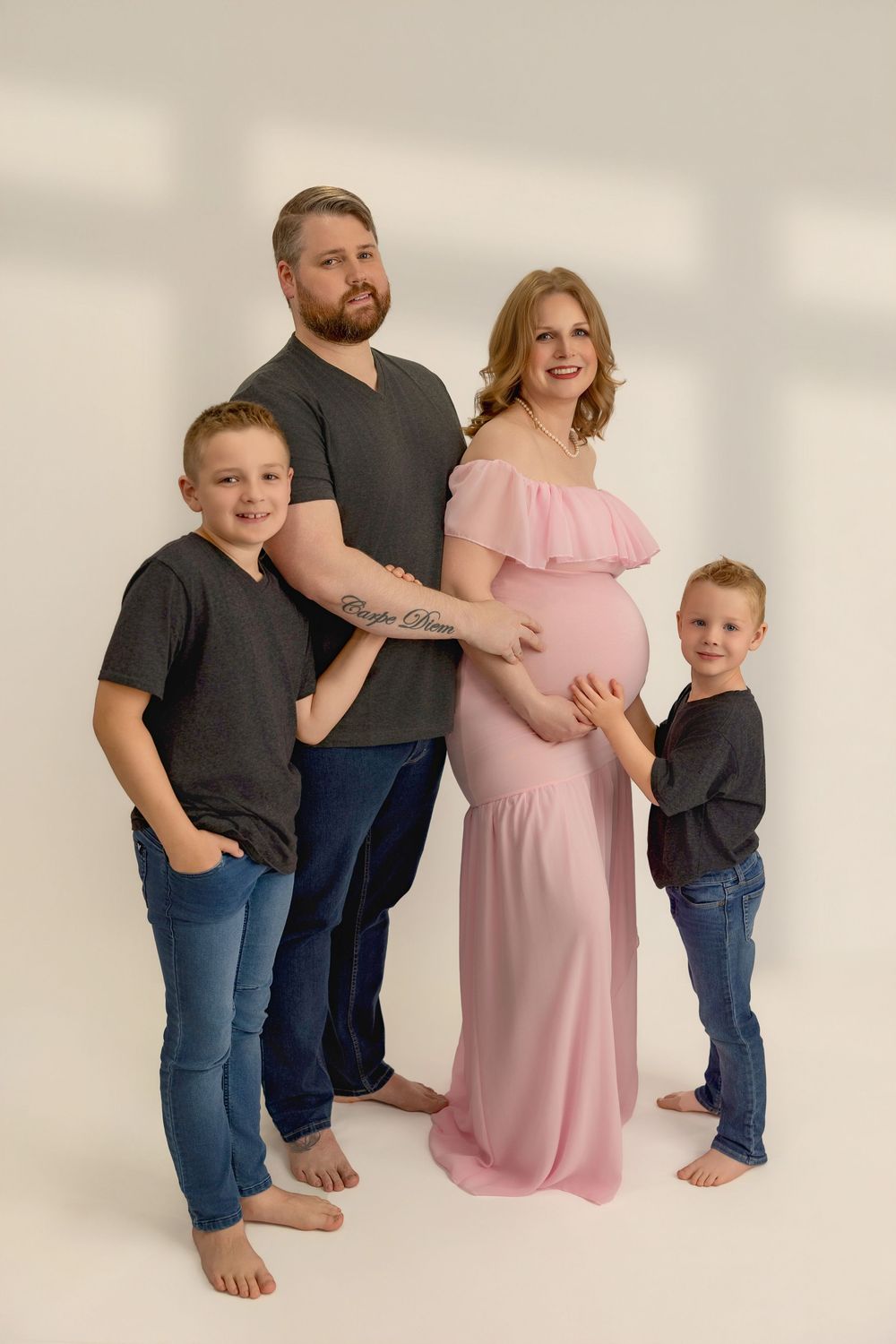 Family portrait with two children in dark shirts and jeans, and an expectant mother wearing a pink off-shoulder maternity gown.