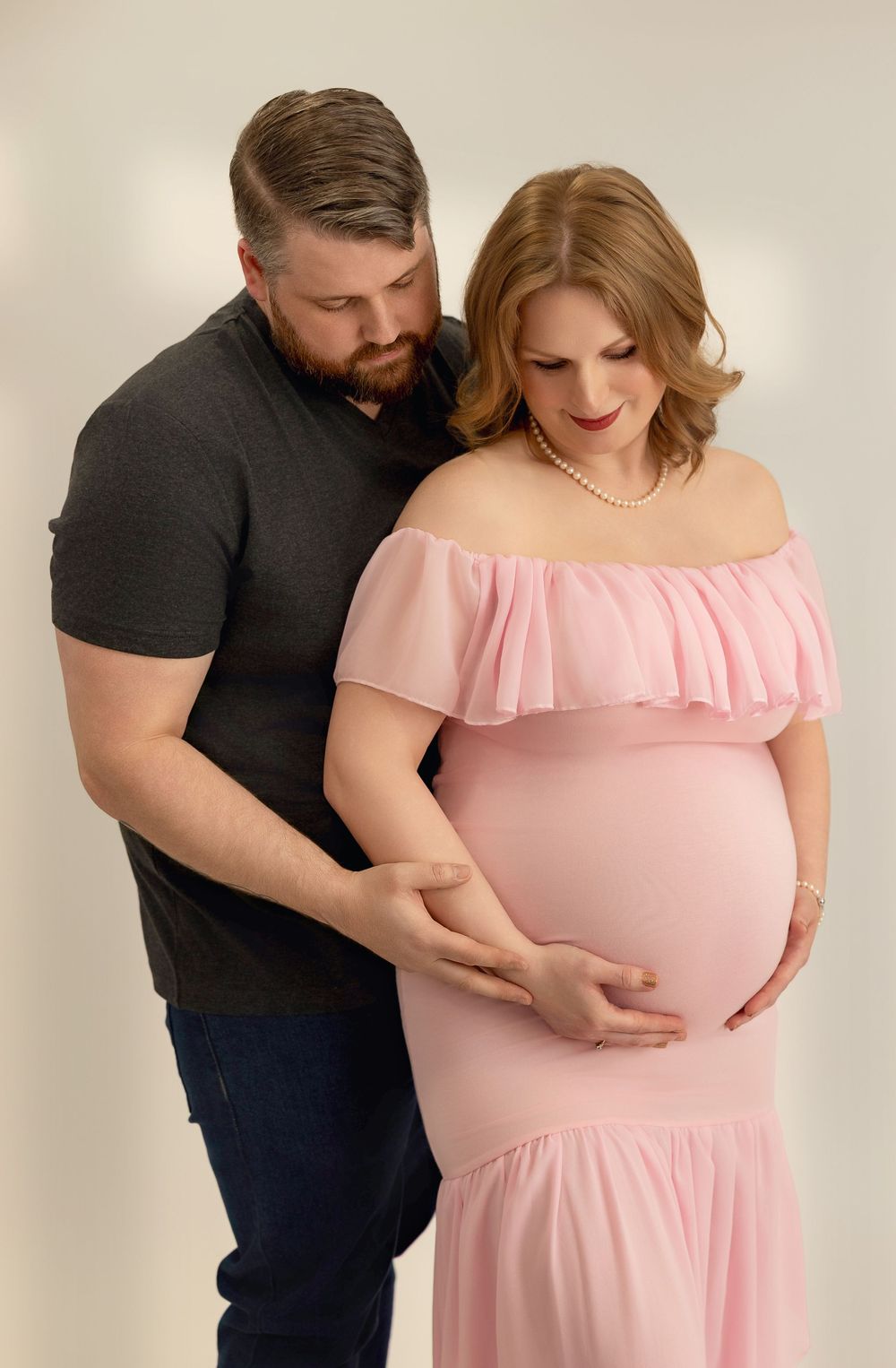 A couple embraces in a maternity photo, with the pregnant person wearing a pink off-shoulder dress against a neutral backdrop.