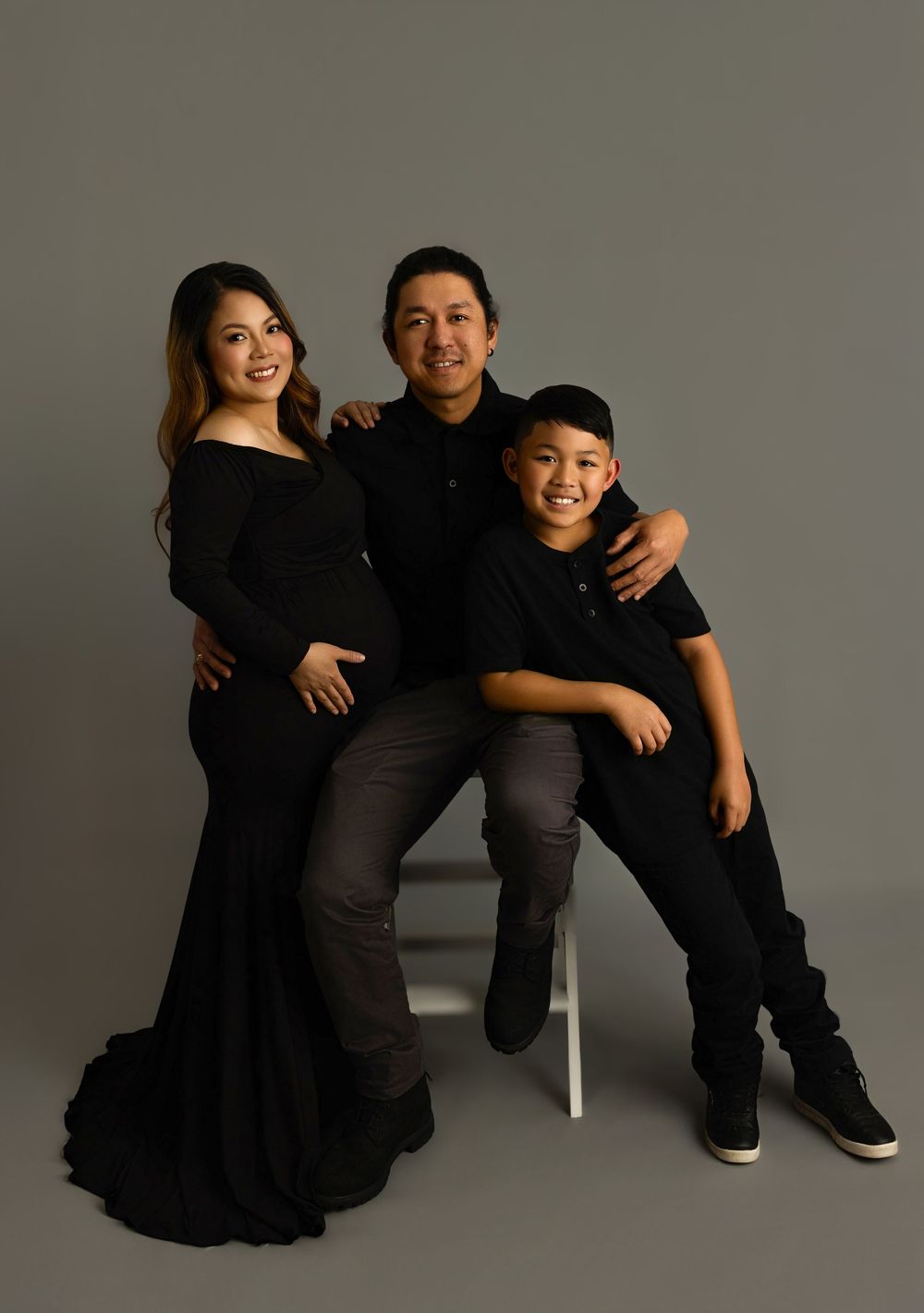 Two figures in black attire pose together in a relaxed studio setting against a gray backdrop.