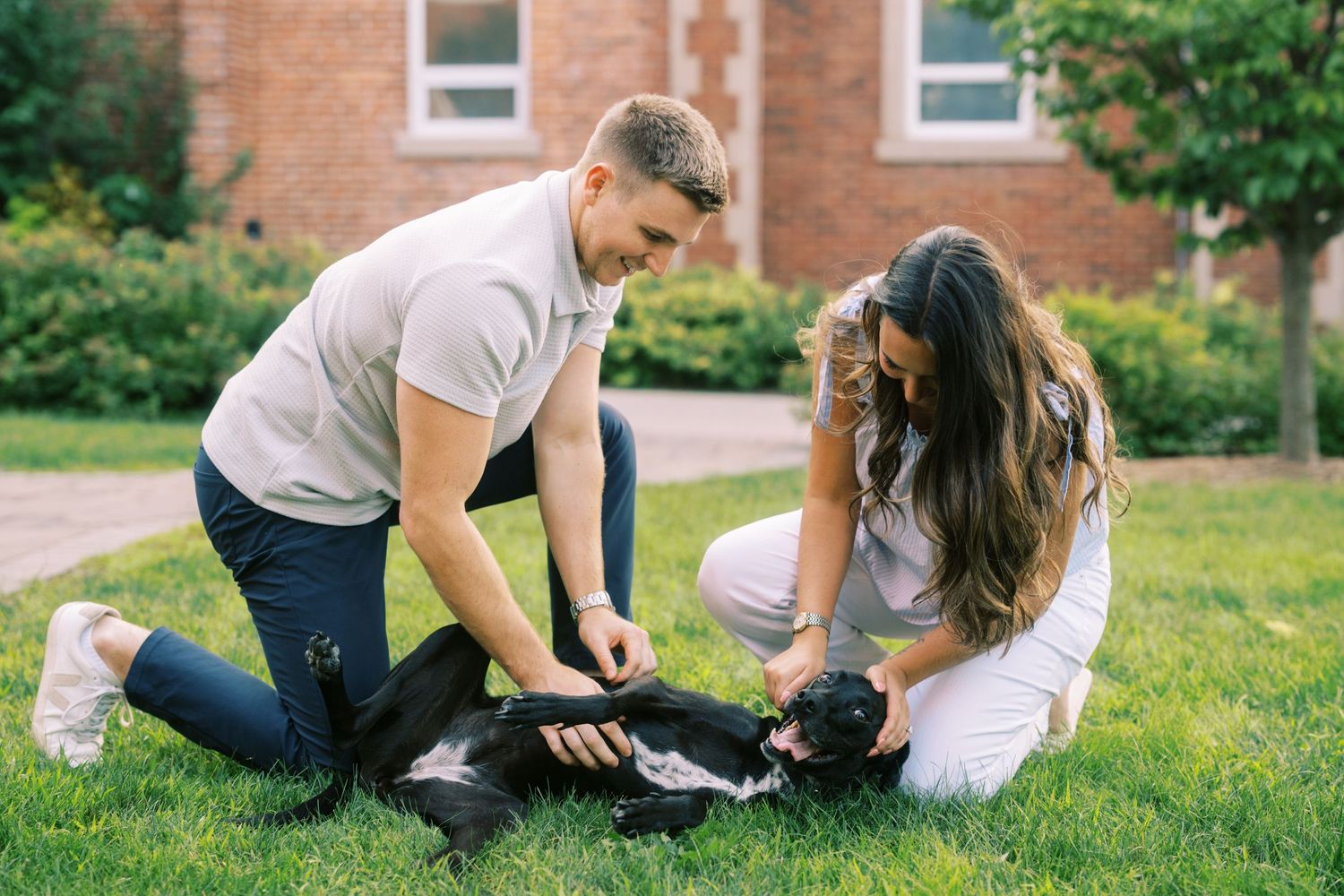Two people kneel on grass while petting a black dog together on a summer day near a brick building.