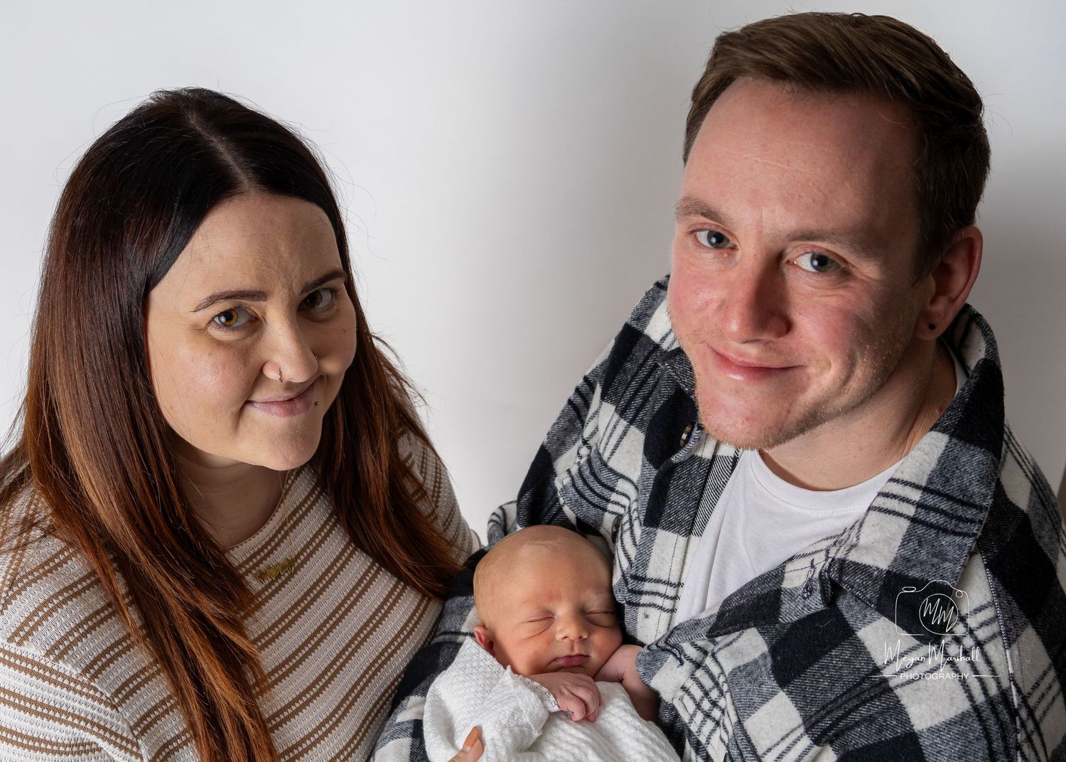 Newborn Photography - A couple in casual sweaters holds their newborn baby against a light gray studio backdrop.