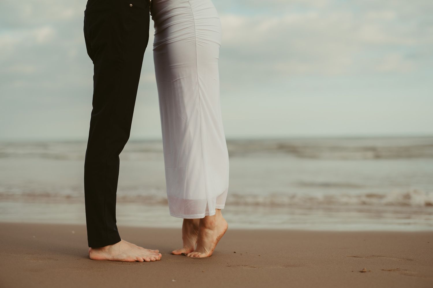 Barefoot couple standing close together on a sandy beach with ocean waves in the background.