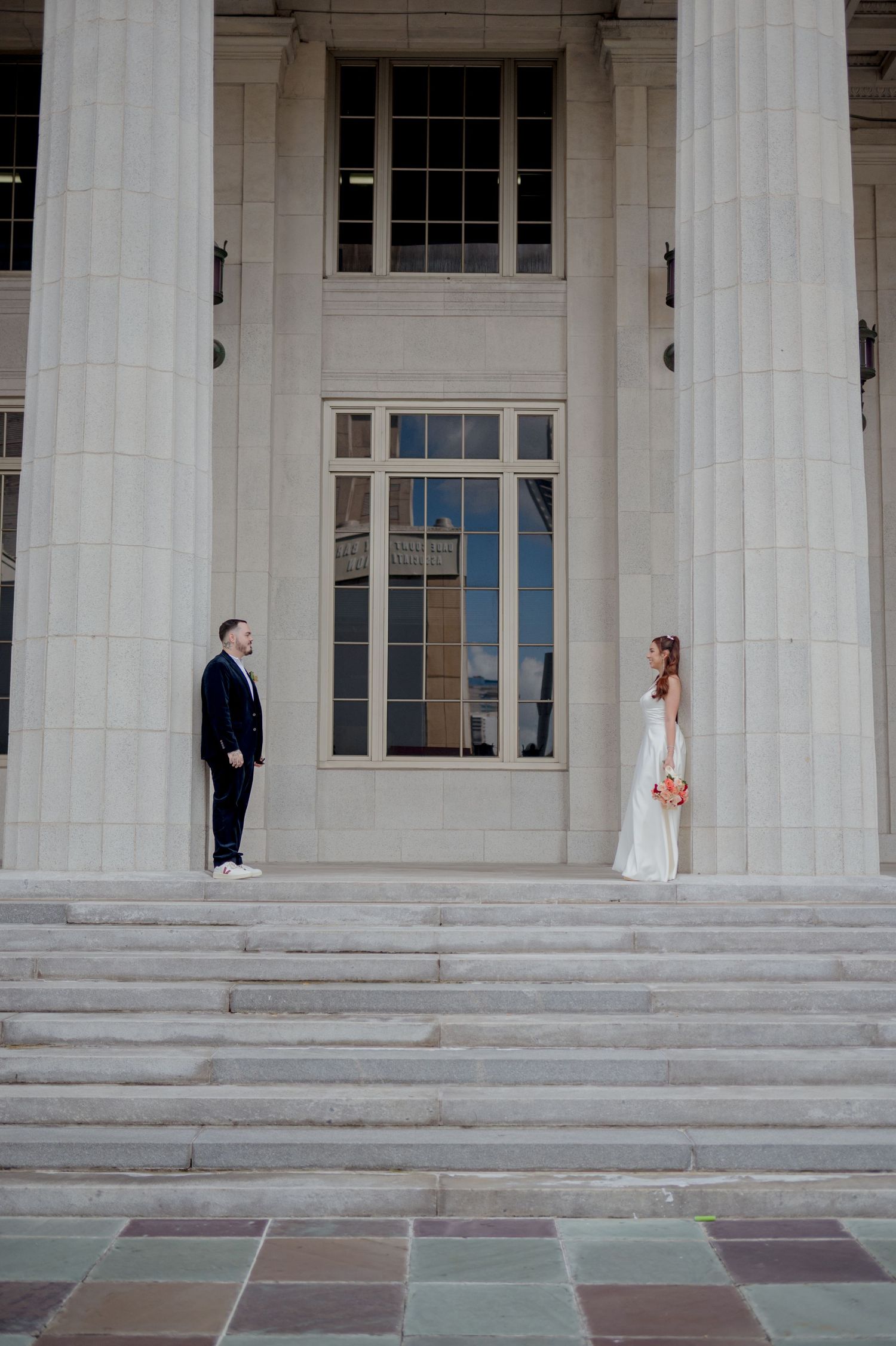 A wedding couple poses separately between elegant white columns of a grand building with stone steps.