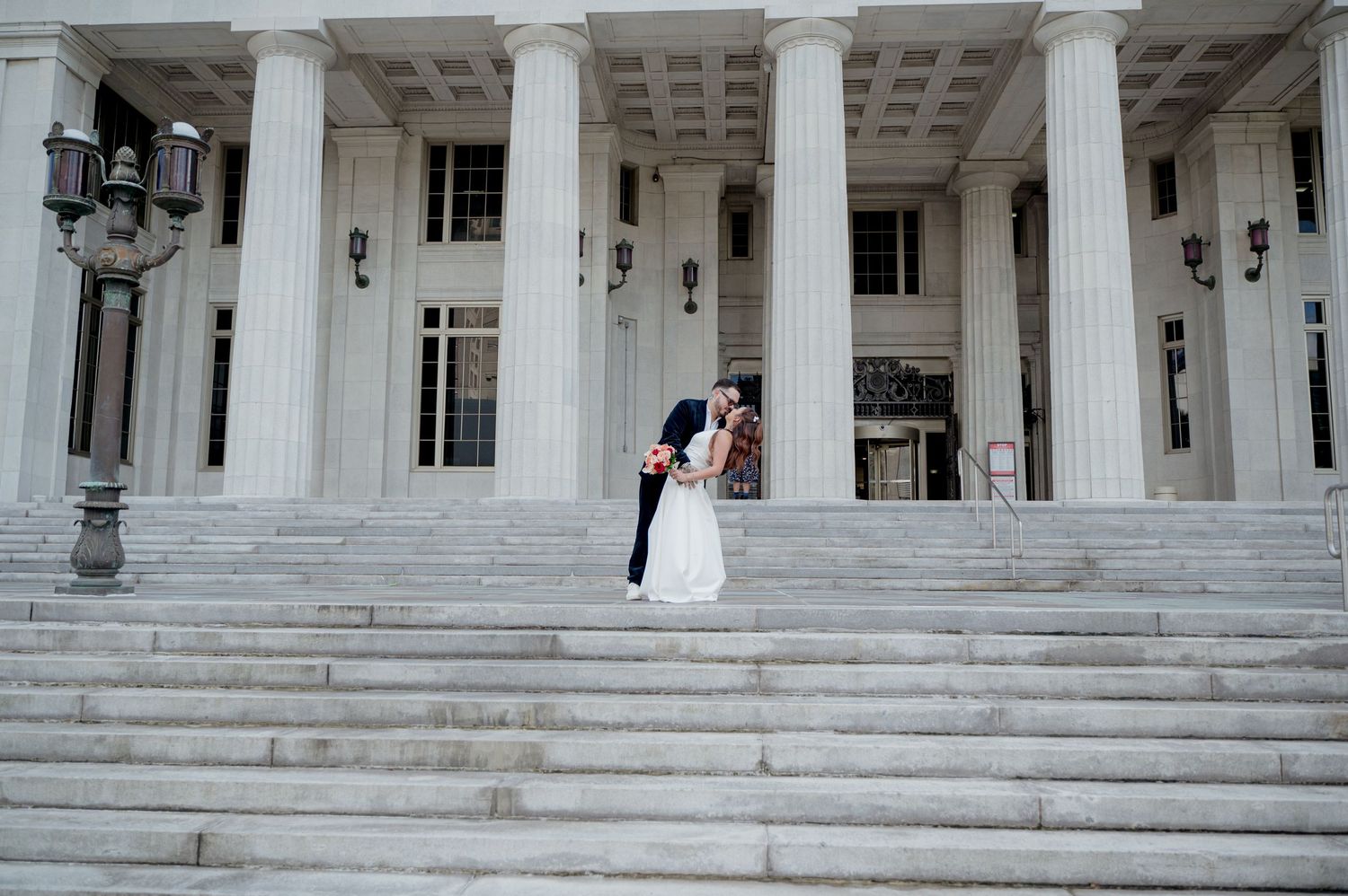 A wedding couple stands on grand white marble steps of a historic courthouse building with elegant columns.