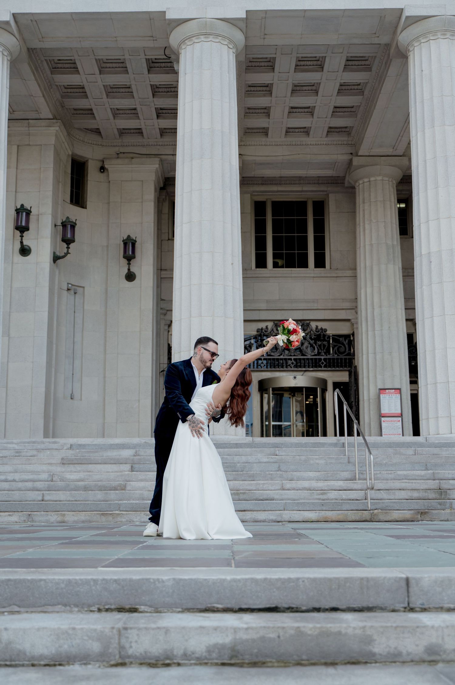 A wedding couple performs a romantic dip pose on marble steps in front of a grand building with classical columns.