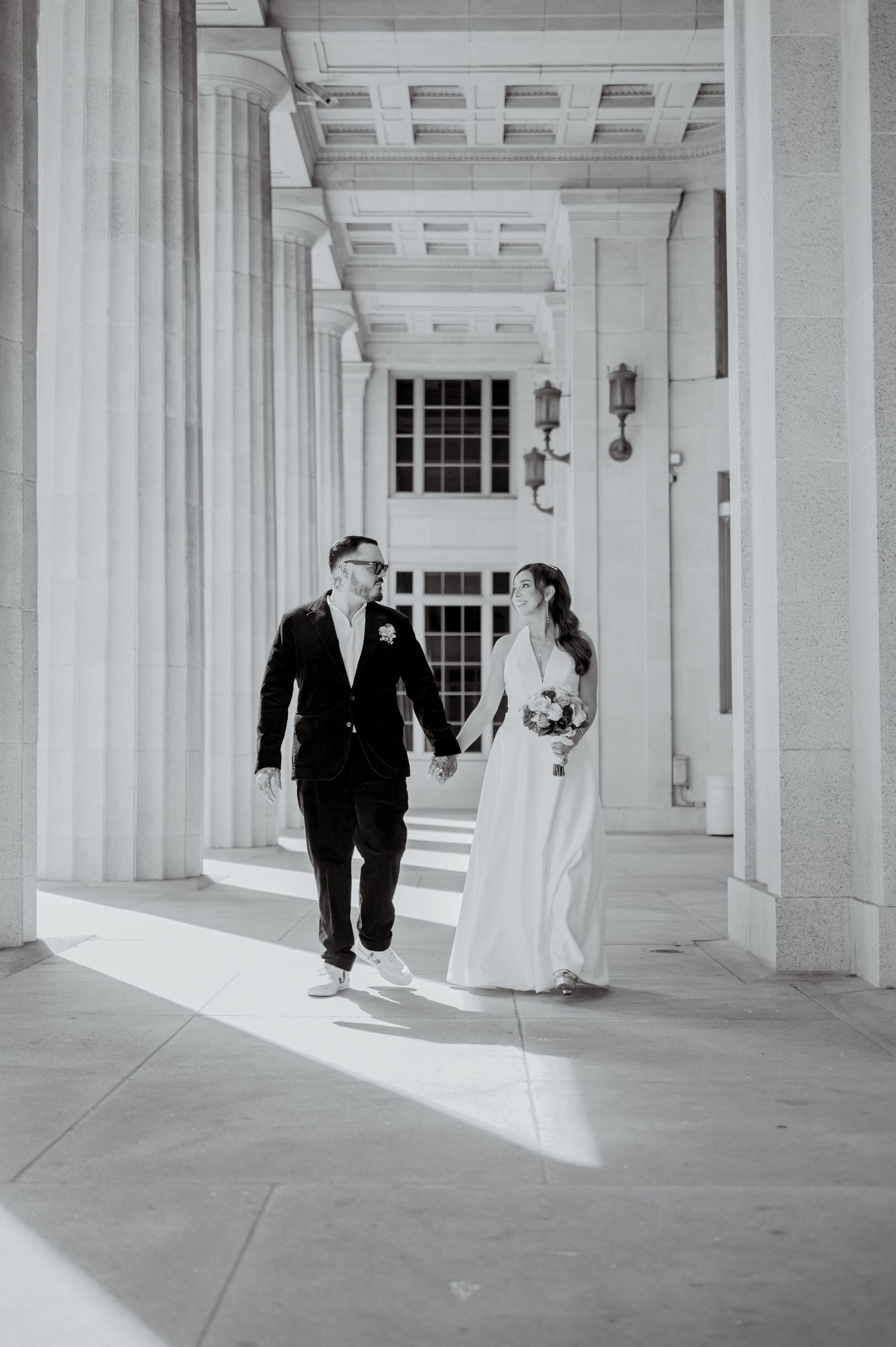 A bride and groom walk through a colonnade of white pillars in a classical architectural setting.
