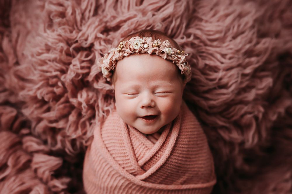 A sleeping newborn baby wearing a pink knit wrap lies on a soft pink fur blanket with a delicate floral crown.