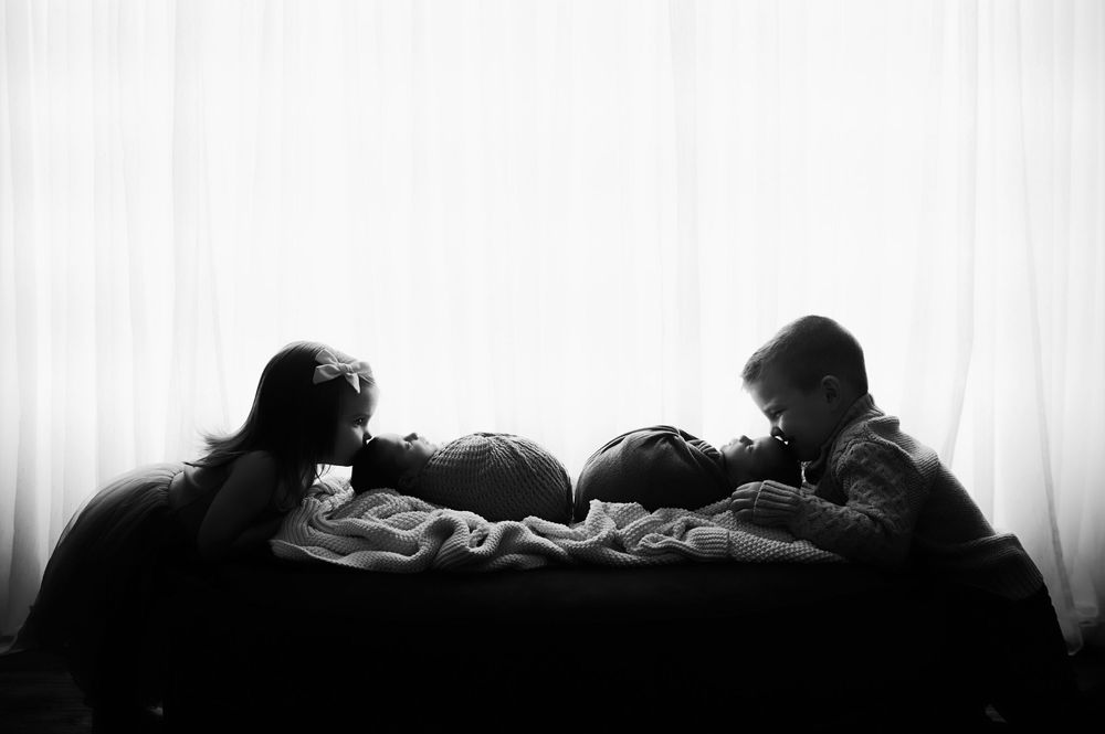 A black and white silhouette of kids snuggling their twin newborn babies.