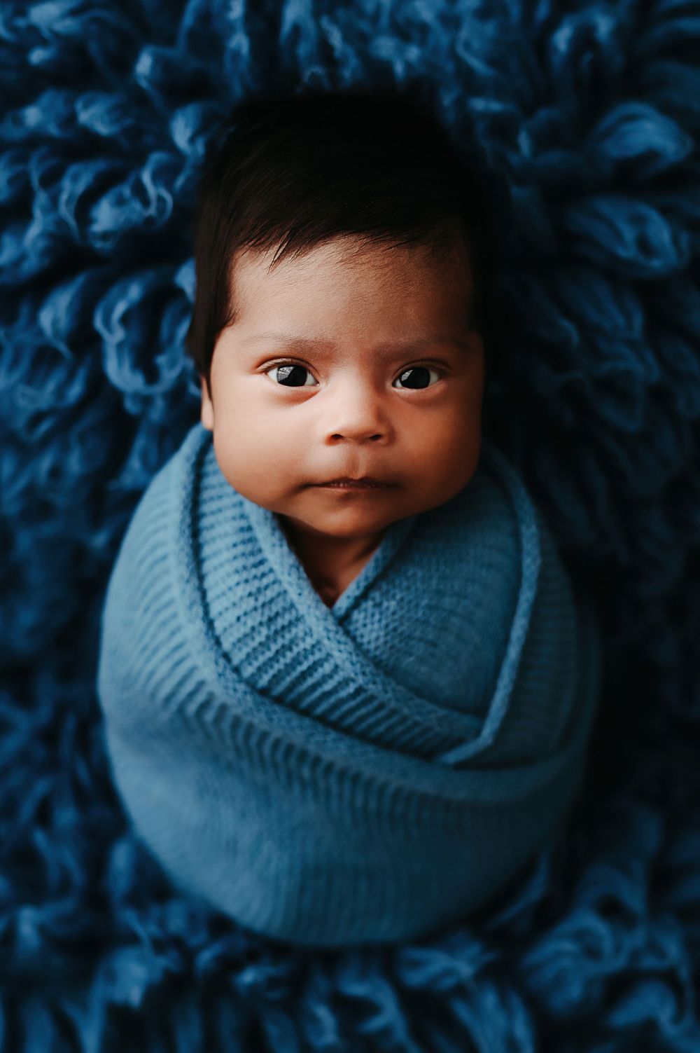 Close-up photos of a baby swaddled in a soft blue knit blanket against a dark blue textured background.