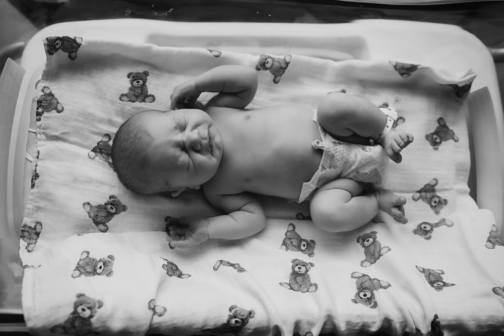Black and white photo of a newborn baby lying on a teddy bear print blanket in a hospital bassinet.