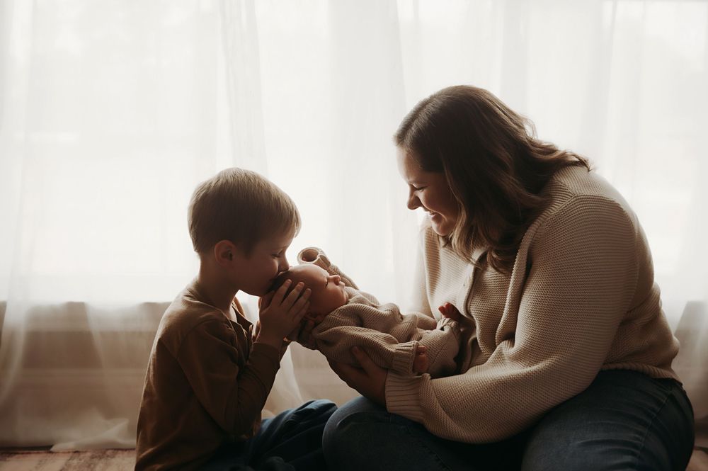 A mother tenderly holds her newborn baby while her young son leans in to meet the infant by a bright window.