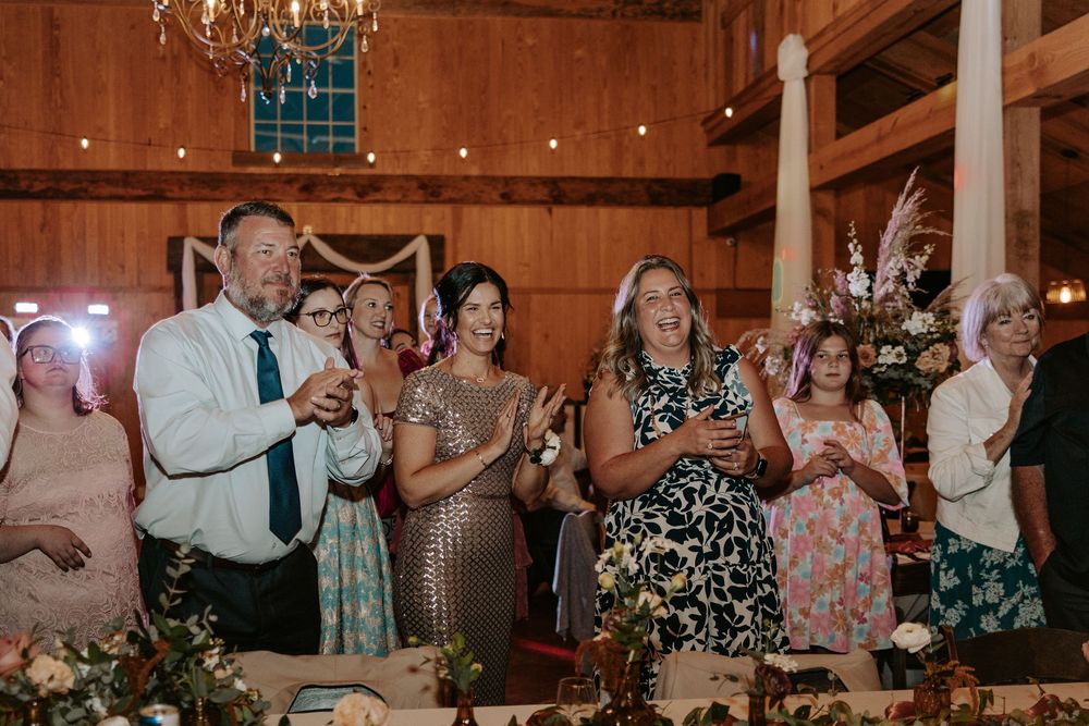 Wedding guests stand and applaud during a reception celebration in a rustic wooden barn venue.