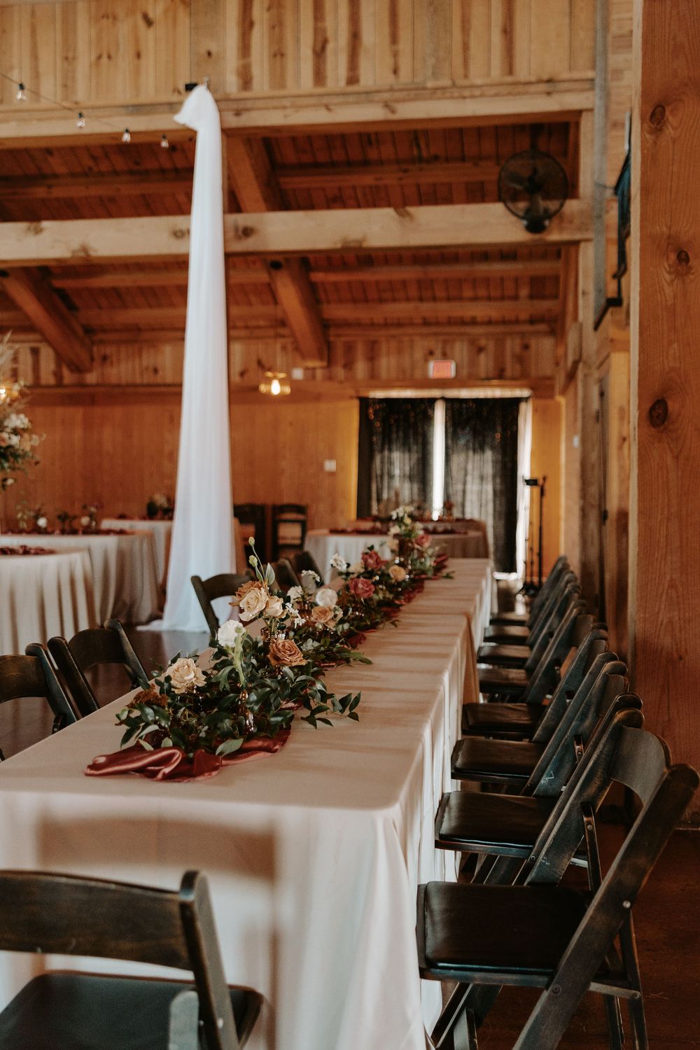 Rustic wedding reception venue with white tablecloths and dark chairs arranged in elegant rows under wooden beams.