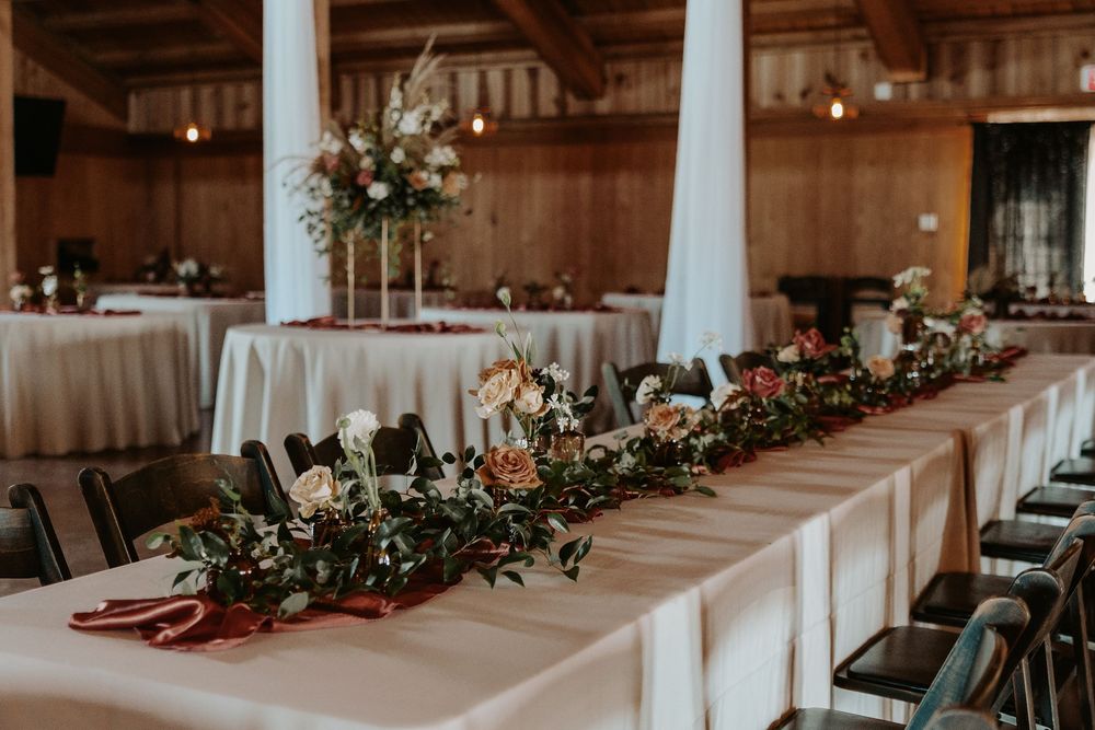 Elegant wedding reception hall decorated with white linens and greenery garlands along rustic wooden beams.