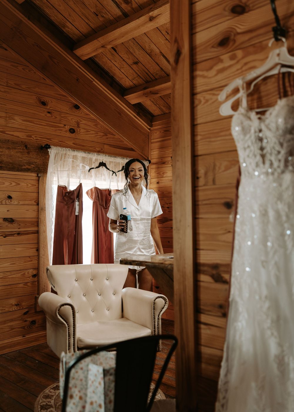 Wedding dresses and robes hanging in a cozy wooden bridal suite with tufted chair.