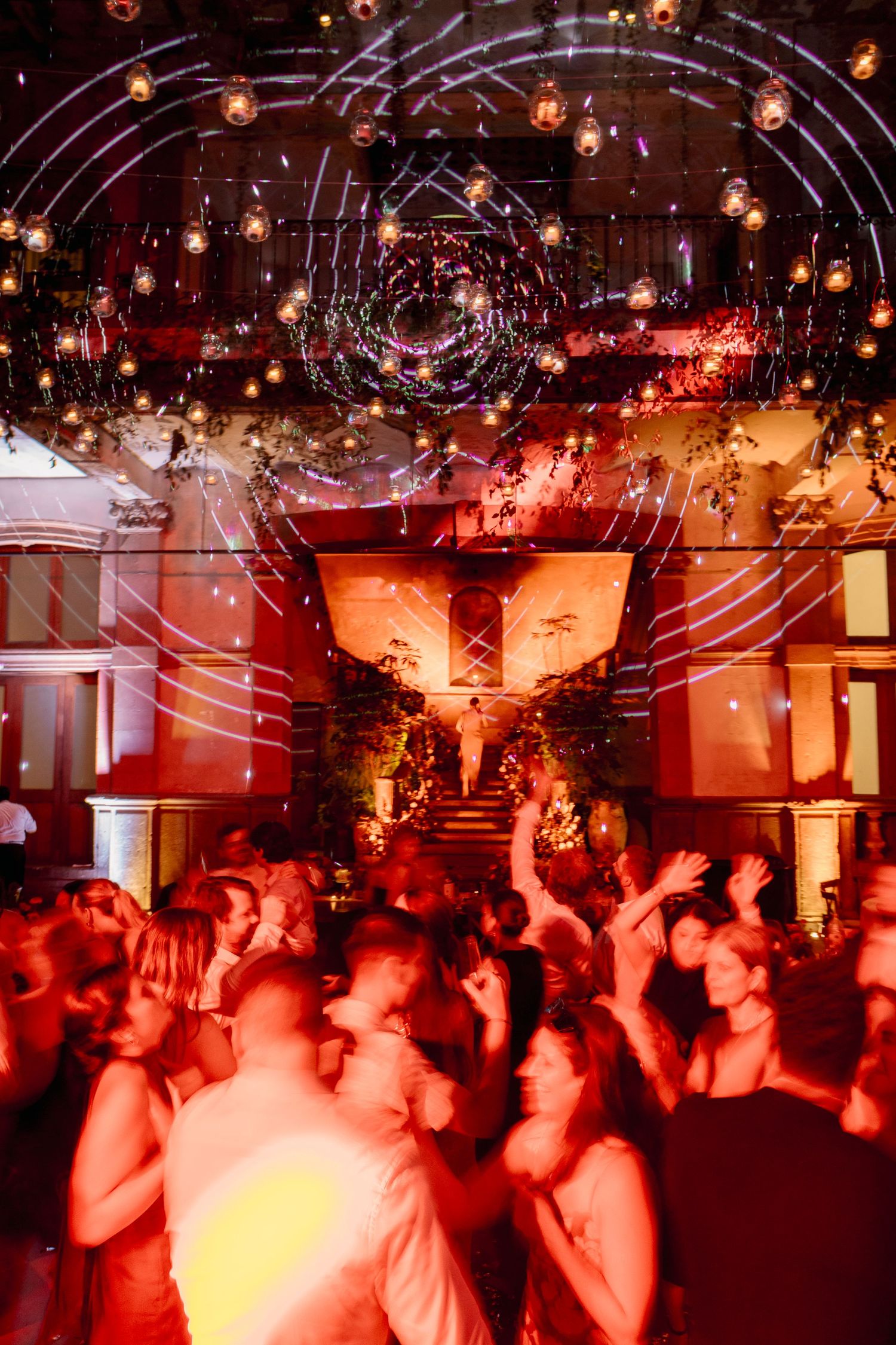A crowded nightclub dance floor illuminated in red lighting with disco balls hanging from the ceiling.