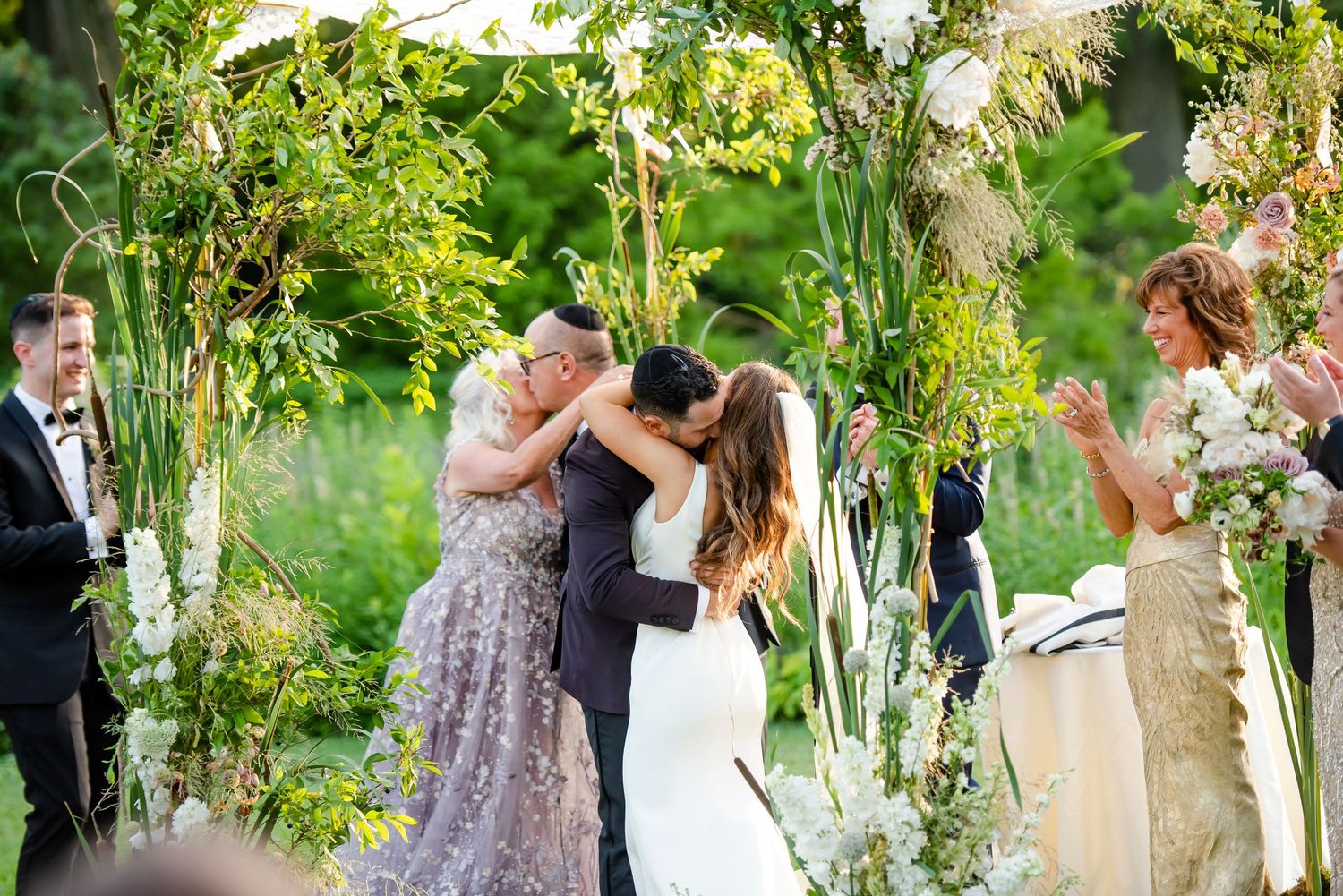 Wedding ceremony under floral arch with couple sharing first kiss while guests celebrate.