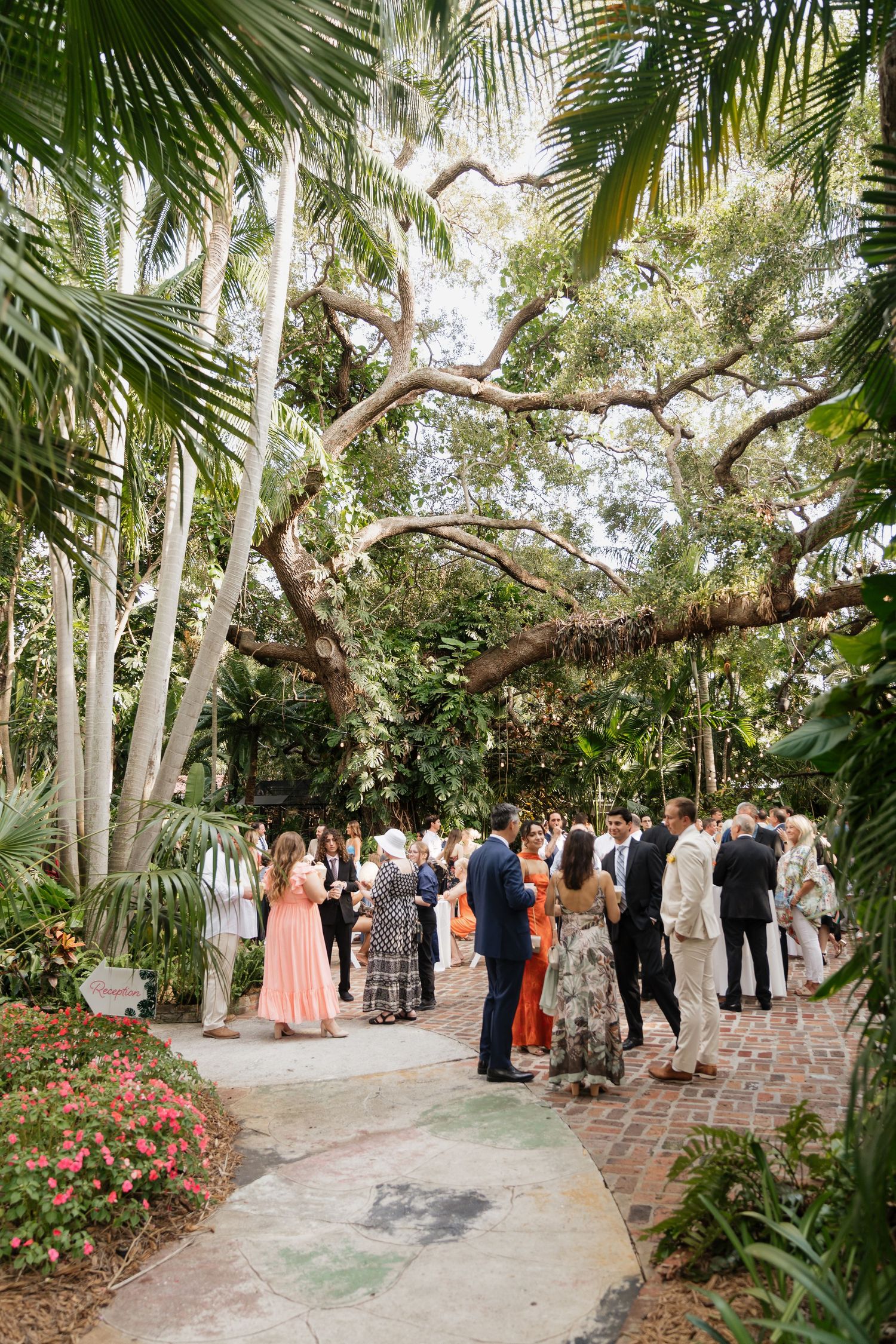Wedding guests gather on a palm-lined brick pathway in a tropical garden setting during an outdoor celebration.