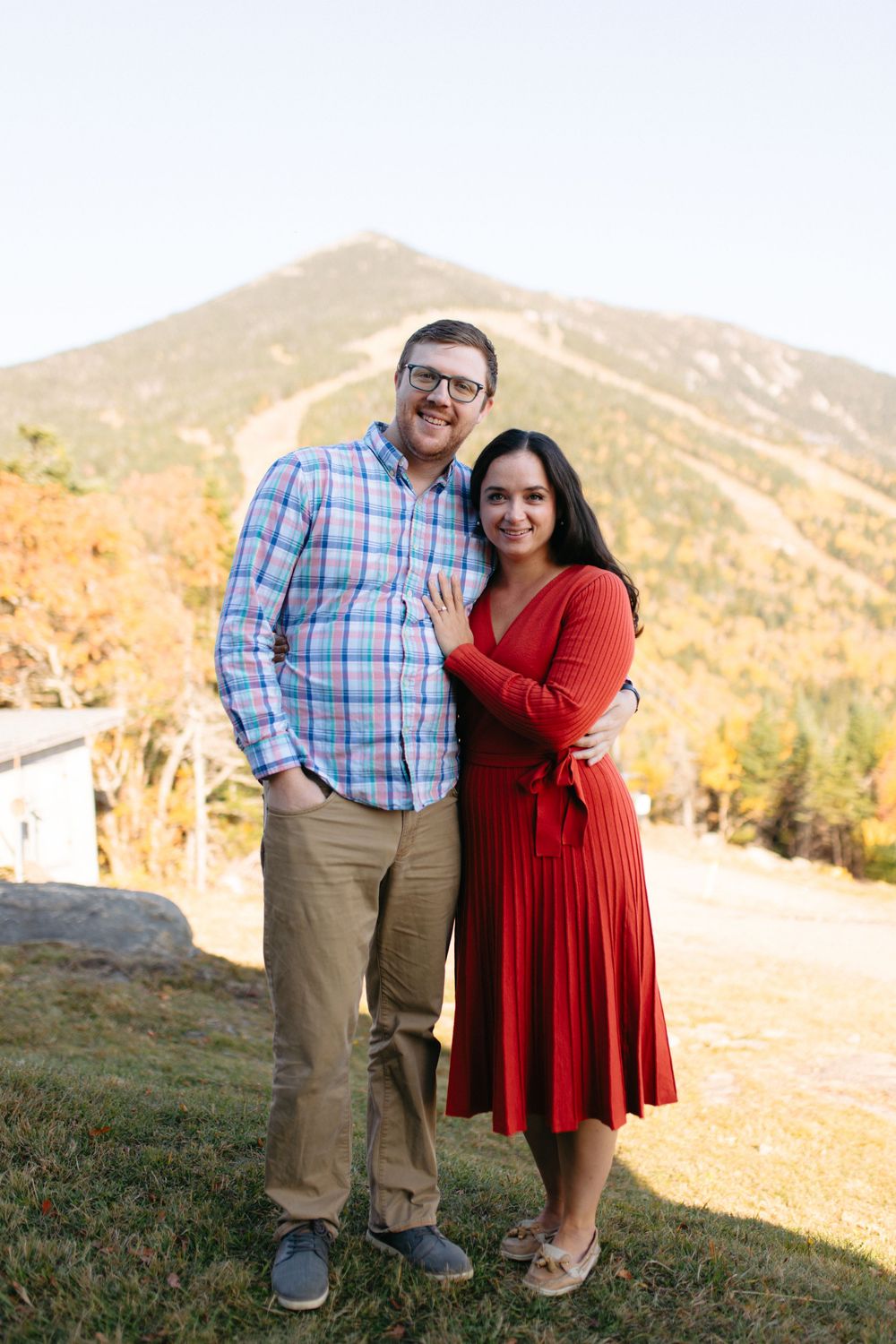 A couple poses together on a hillside during golden hour, with her wearing a red dress and him in a plaid shirt.