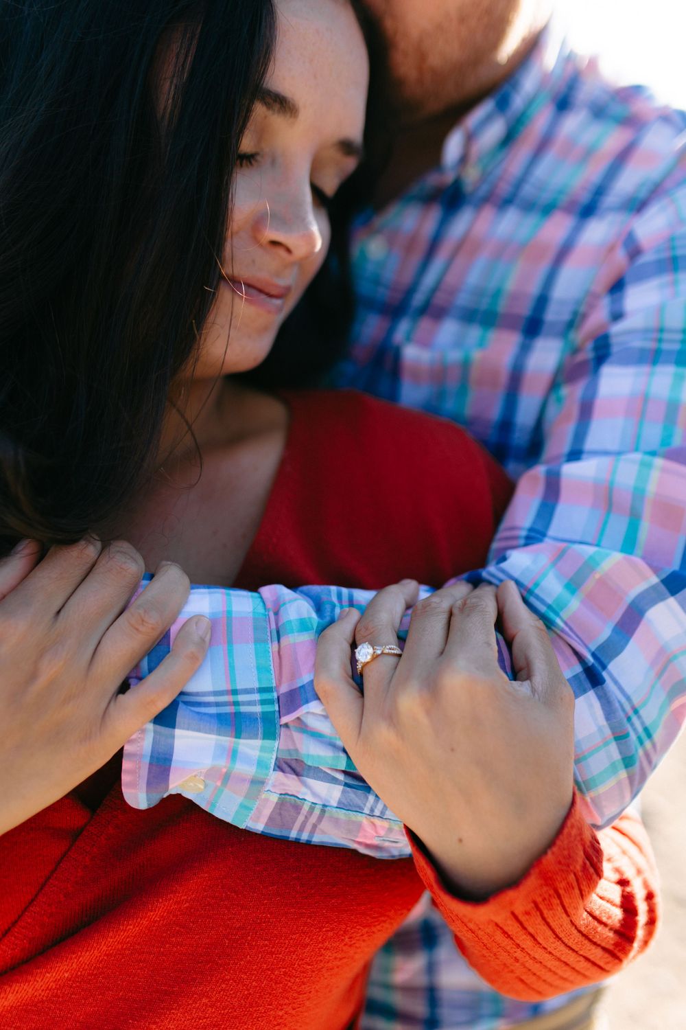 A couple embraces tenderly while wearing a red sweater and plaid shirt, showing off an engagement ring.