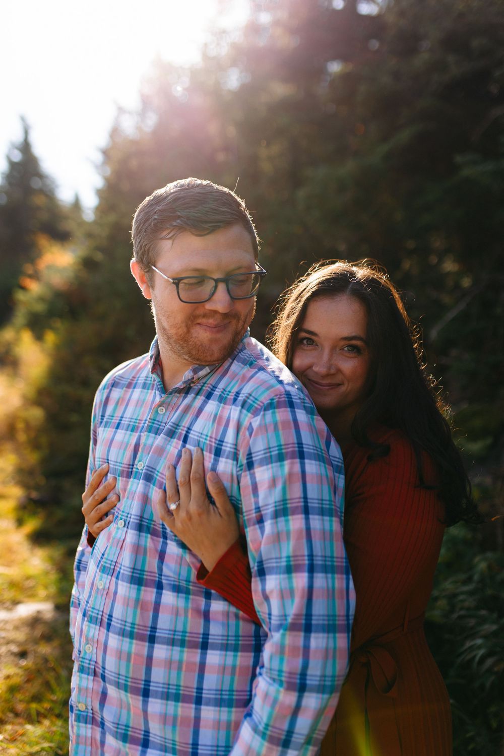 A romantic outdoor engagement photo with a couple in matching red and plaid against a sunlit forest backdrop.
