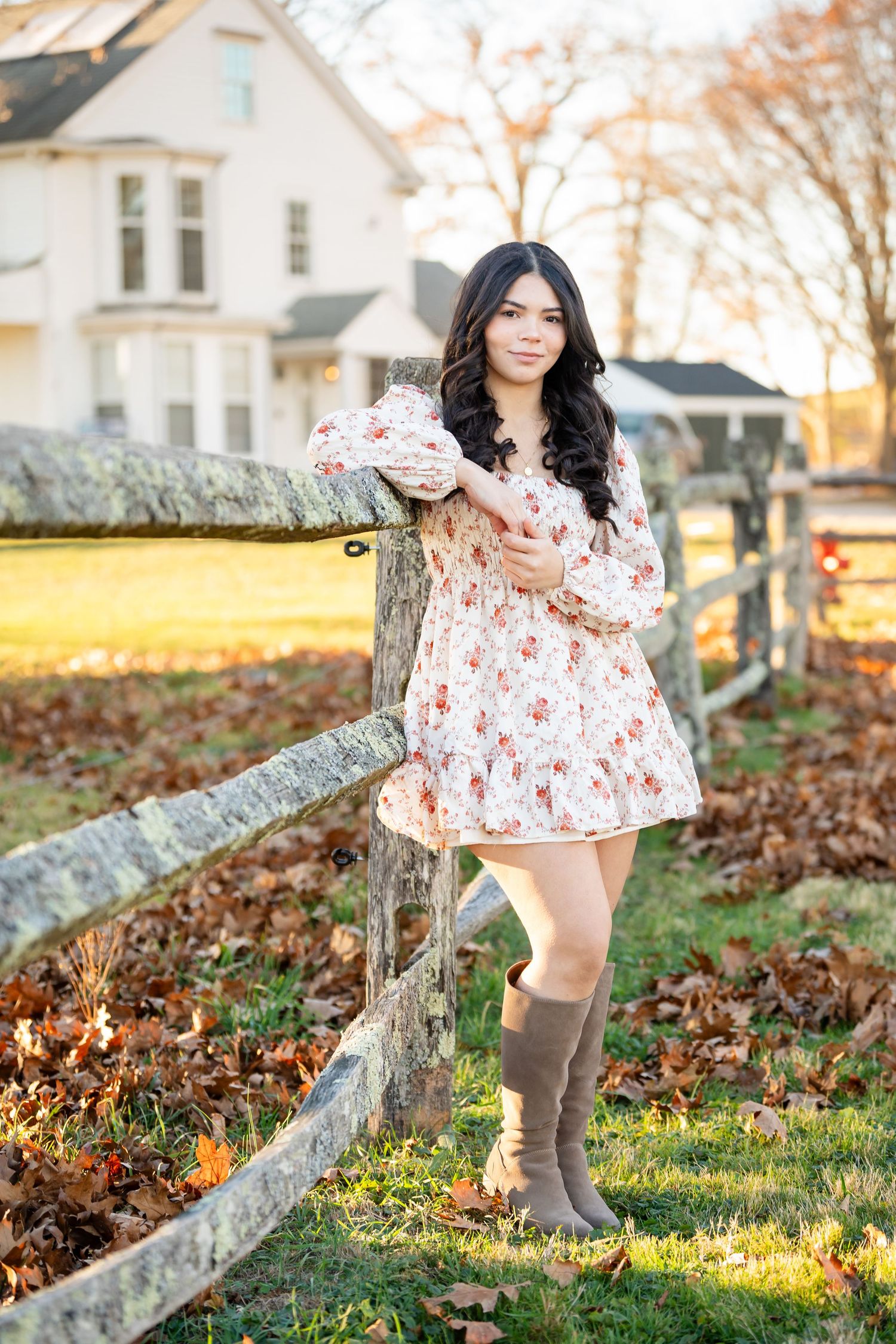 A person in a floral dress and brown boots poses by a rustic wooden fence during autumn sunset.