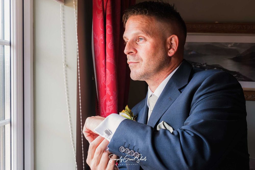 A photo of the groom looking out of a window whilst adjusting his cufflink, captured by a Wedding Photographer at Winters Tale Country Barn