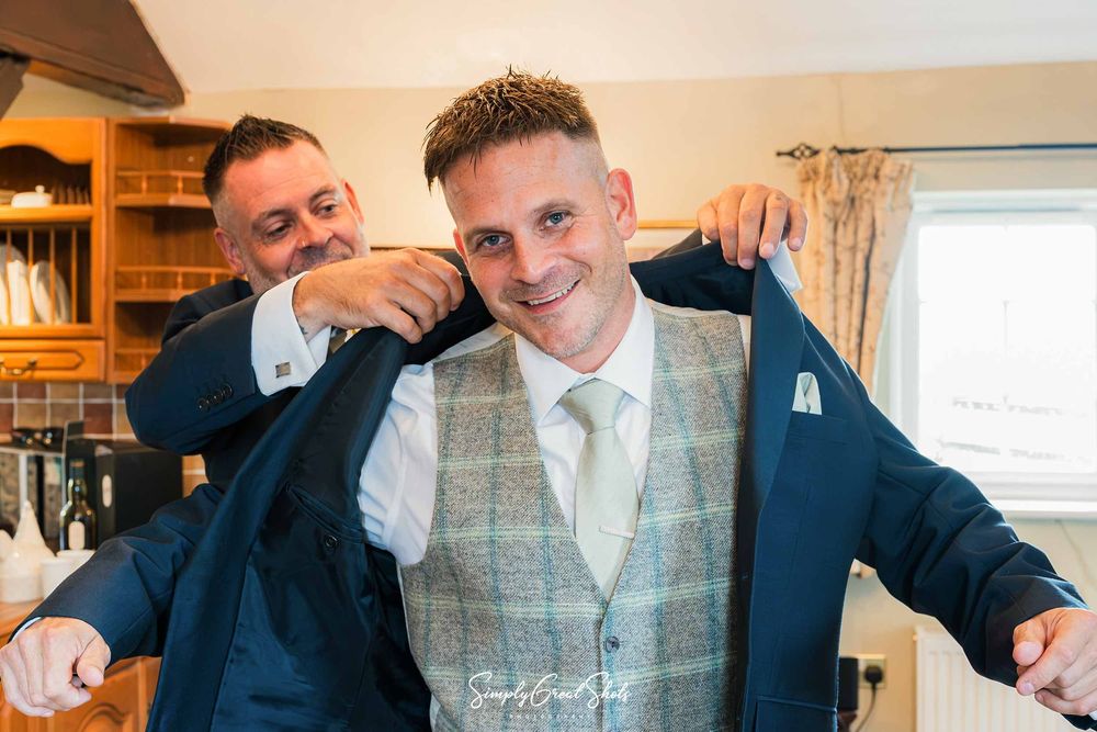 In the Ember Eve Barn, the Groom gets a helping hand from his Best Man putting on his jacket over a Sage and Blue Harris checked waistcoat