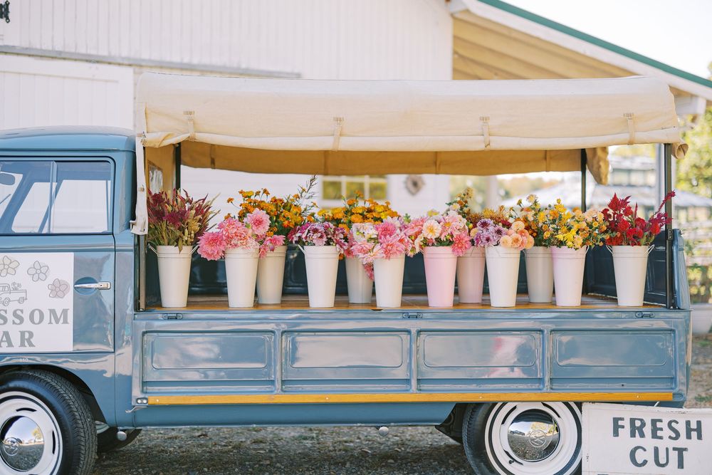 A vintage blue flower truck displays colorful blooms in pink ceramic pots along its wooden bed at a farmers market.