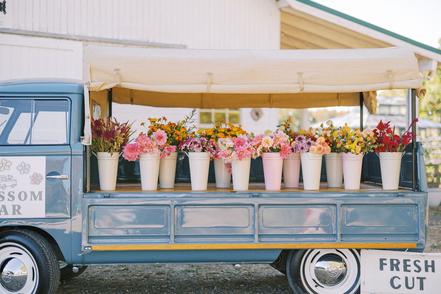 A vintage blue flower truck displays colorful blooms in pink ceramic pots along its wooden bed at a farmers market.