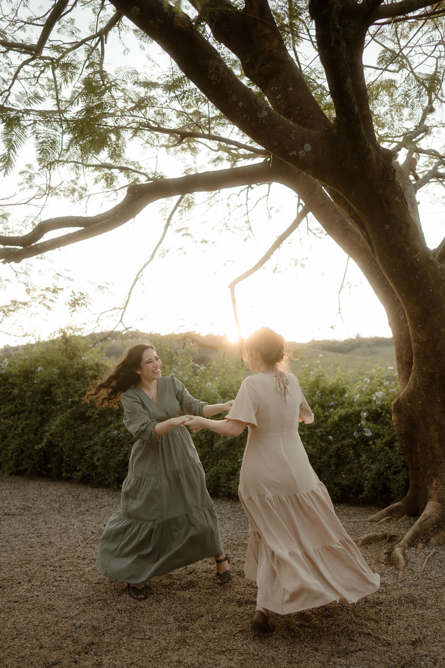 Two figures in flowing dresses dance together under a sprawling tree at sunset creating a dreamy atmosphere.