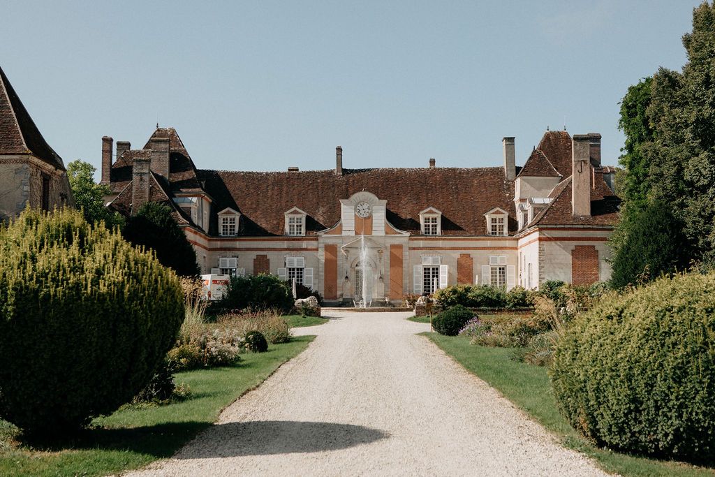 Elegant French chateau wedding venue with a tree-lined gravel driveway under a clear blue sky.