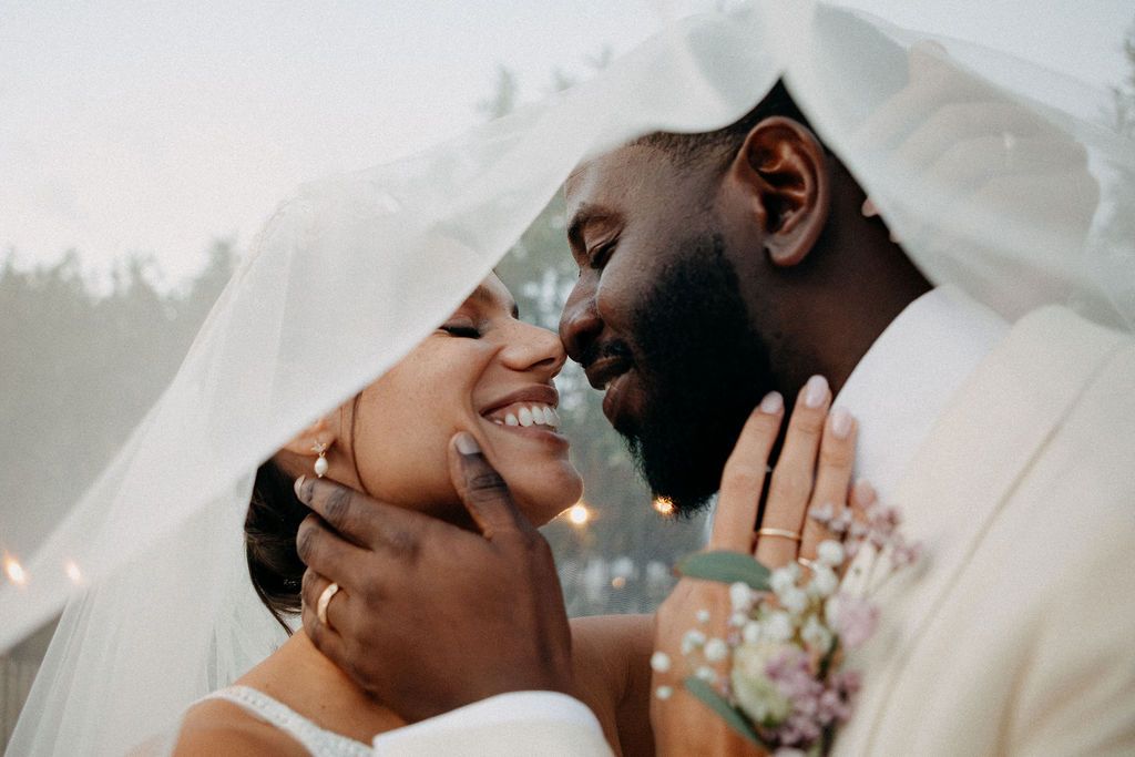 Romantic interracial bride and groom sharing an intimate moment under a white veil at their wedding.