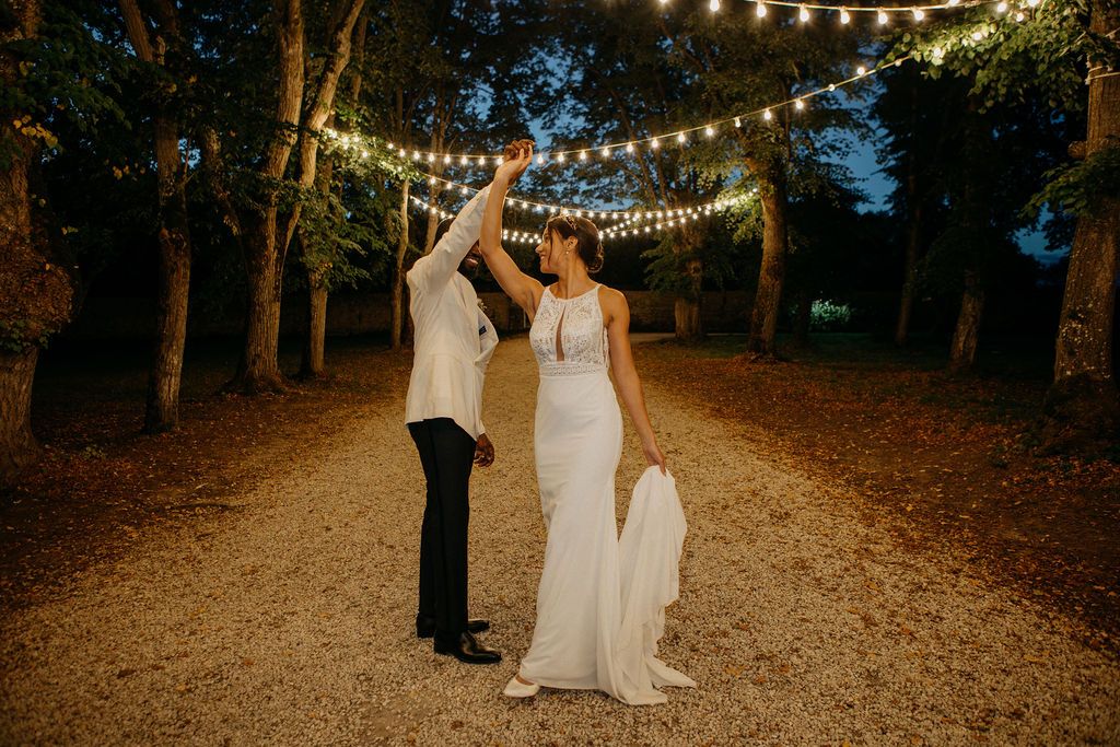 Bride and groom dancing under string lights on a tree-lined path at night during wedding celebration.