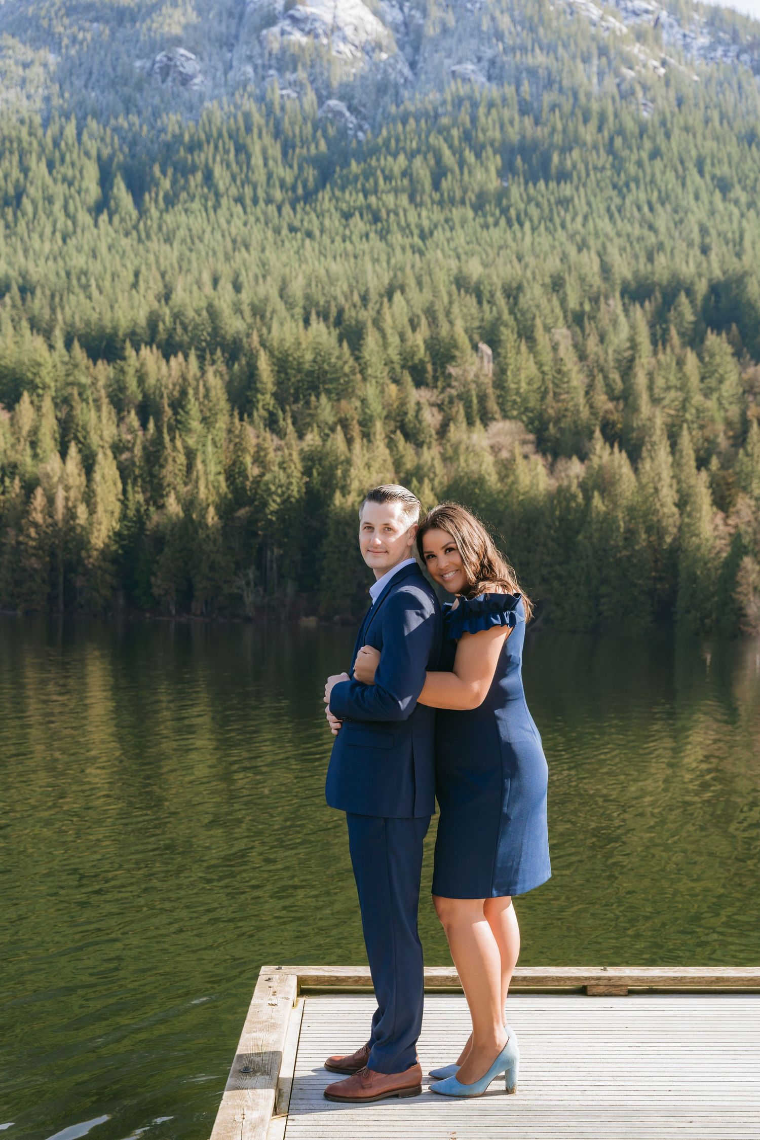 Two people with umbrellas walk along a wooden dock on a misty lake surrounded by evergreen-covered mountains.