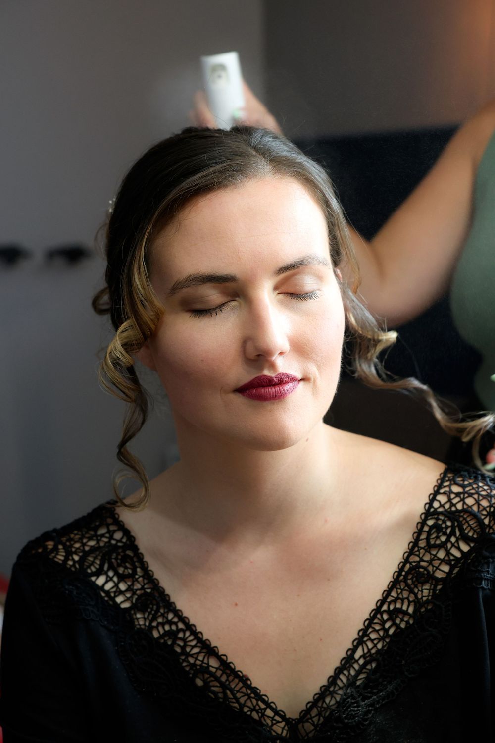 A person wearing a black lace top with red lipstick and curled hair looks down with closed eyes.