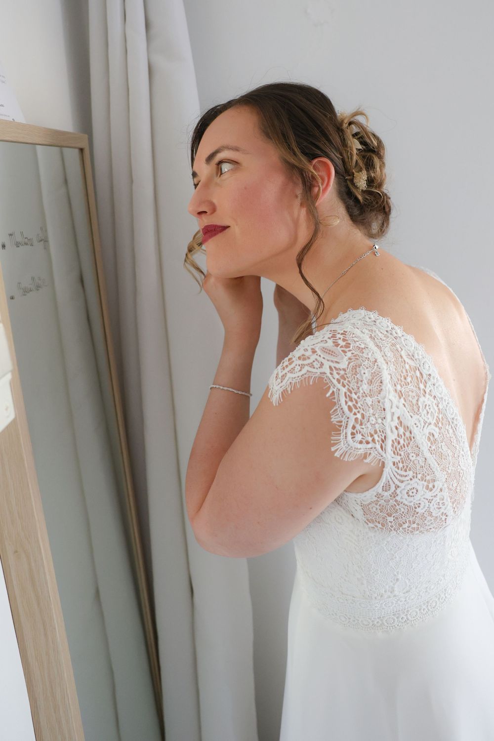 A bride in an elegant white lace wedding dress with sheer back detail gazes thoughtfully by a window.