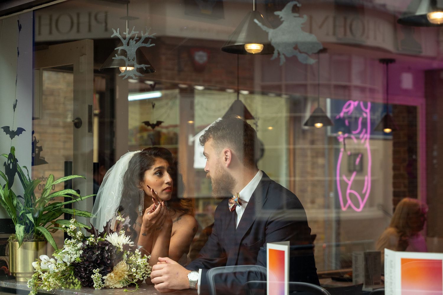 Couple sits at restaurant table near window with floral bouquet and neon lights glowing in background.
