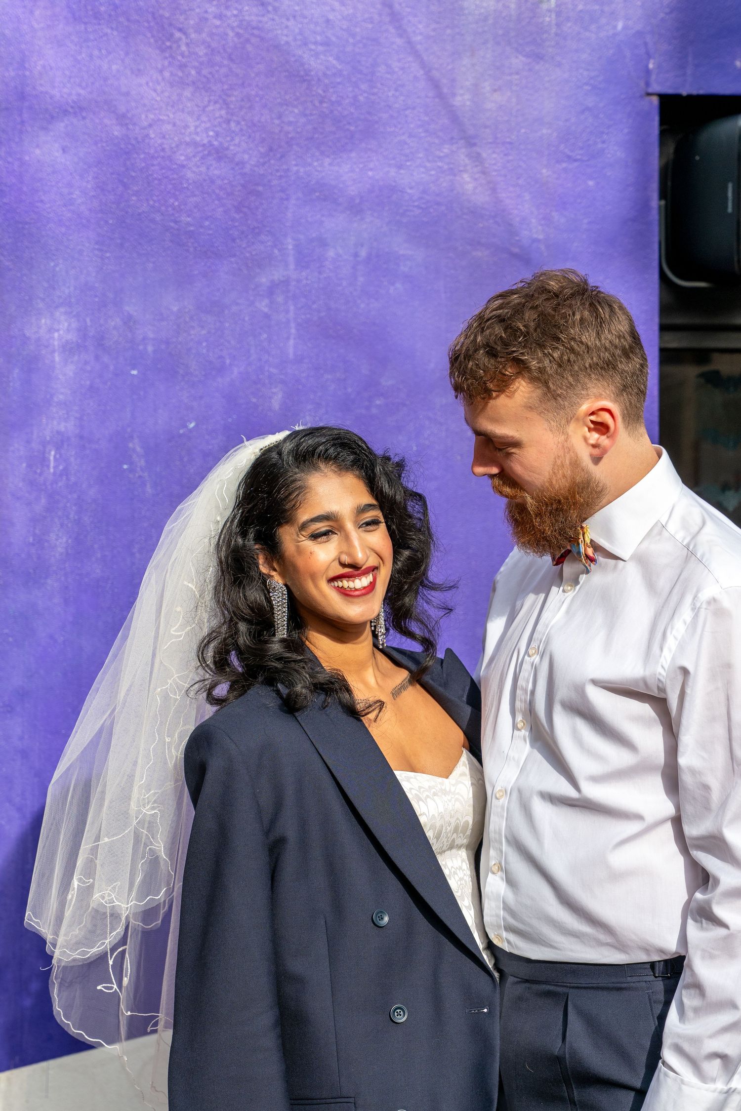 A couple shares a joyful moment against a vibrant purple wall during their wedding photoshoot.