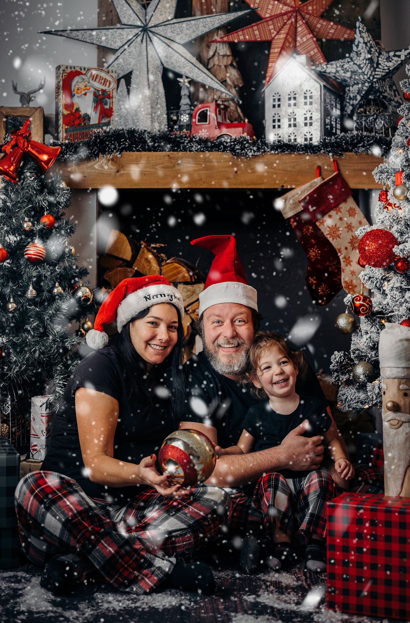 Family wearing Santa hats and plaid pajamas pose together amid falling snow and Christmas decorations.
