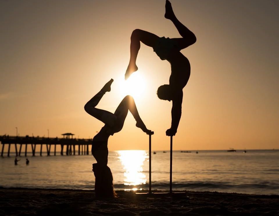 Silhouettes of gymnasts performing acrobatic moves at sunset by the beach.