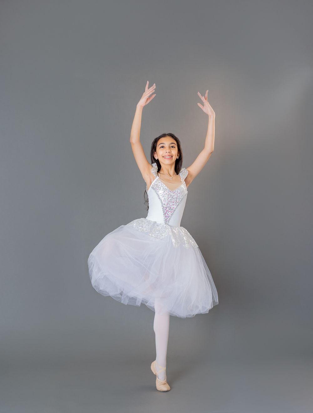 Young ballet dancer in white tutu dress performs graceful dance poses against a gray studio background.